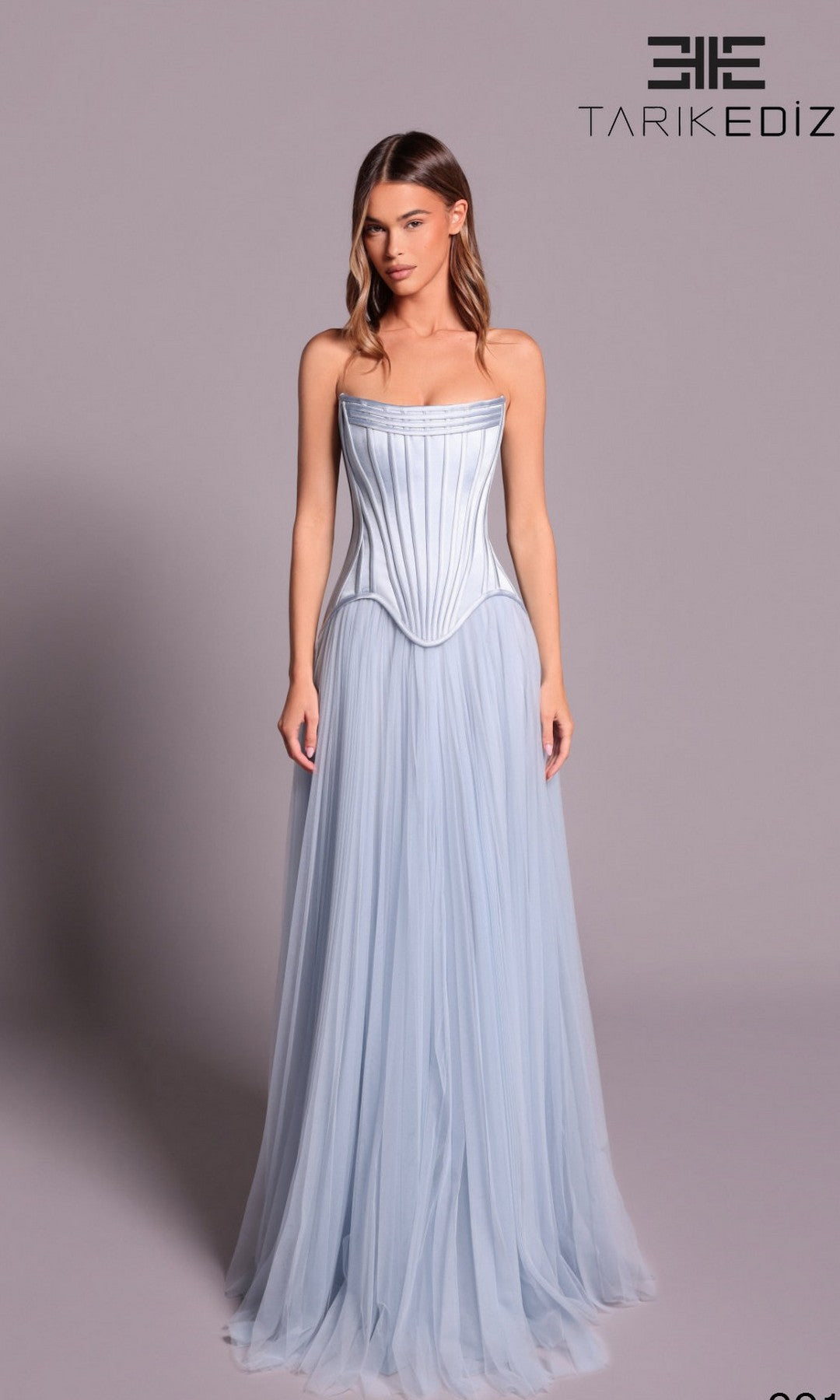 A woman wears the Tarik Ediz 99137 Long Formal Dress in Blue Aura, featuring a strapless corset bodice and flowing tulle skirt. The Tarik Ediz logo appears in the upper right corner.